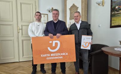 Burmistrz Dzierżoniowa i dwóch przedsiębiorców trzymają dużą pomarańczową planszę z napisem Karta Mieszkańca Dzierżoniowa.