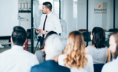 Mężczyzna prowadzi szkolenie w sali konferencyjnej, stojąc przy tablicy i trzymając tablet, a przed nim siedzi grupa uczestników; w tle widoczne logo „Invest in Dzierżoniów”