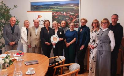 grupa członków Dzierżoniowskiej Rady Przedsiębiorców podczas spotkania w Urzędzie Miasta. Dziewięć osób stoi przy stole konferencyjnym, na tle panoramy Dzierżoniowa i baneru „Invest Dzierżoniów”. Pośrodku jedna z uczestniczek trzyma okrągły znak Rady Przedsiębiorców.