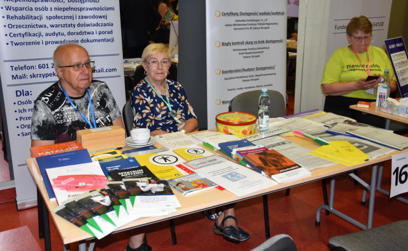 Przy stole siedzi kobieta i mężczyzna. Na stole znajdują się ulotki i książki. Z tyłu stoją roll-upy firmy Kancelaria PRO-3R.