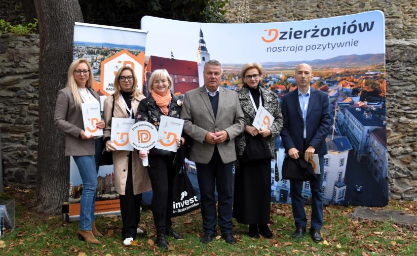 Członkowie Dzierżoniowskiej Rady Przedsiębiorców stoja z burmistrzem przed ścianką promocyjną Dzierzoniowa. 
