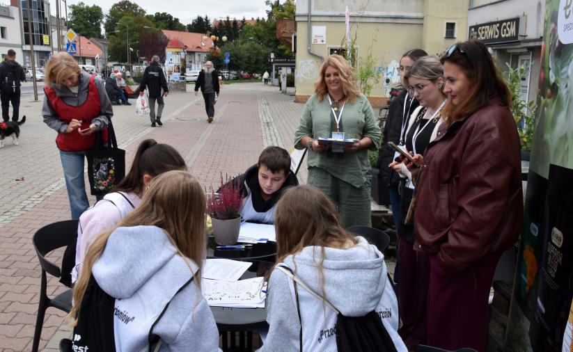 Czwórka uczestników gry siedzi przy stole i rozwiązuje zadanie na stacji gry. Obok stoi opiekun drużyny i organizatorzy stacji.