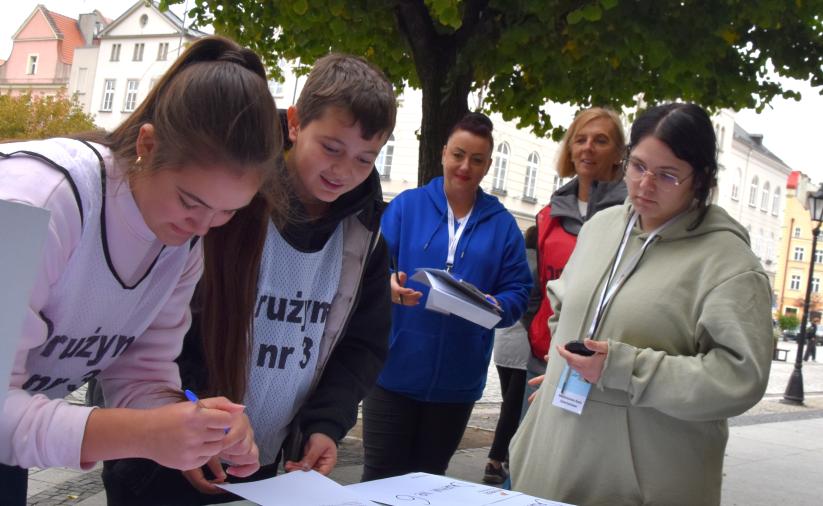 Drużyna pochyla się nad stołem i rozwiązuje zadanie na stacji gry. Obok stoi opiekun drużyny oraz organizatorzy stacji. W tle widoczny jest ratusz miejski w Dzierżoniowie