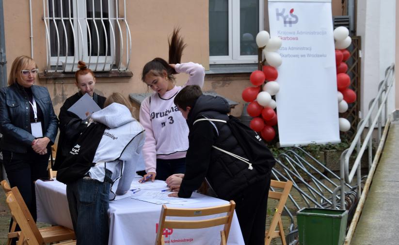 Czworo uczestników gry terenowej rozwiązuje zadanie przy stole nakrytym białym obrusem. W tle stoją dwie opiekunki stacji, biały budynek oraz roll-up z balonami.