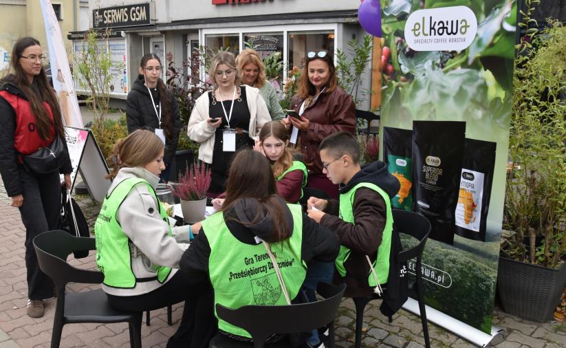 Czwórka uczestników gry pochyla się nad stołem i rozwiązuje zadanie na stacji gry. Obok stoi opiekun drużyny. 