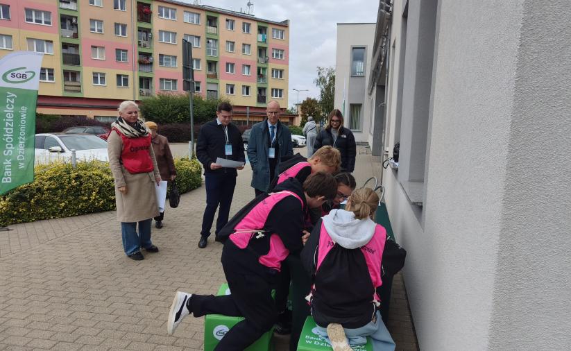 Uczestnicy gry w różowych kamizelkach stoją tyłem, pochyleni nad stołem przy stacji zadaniowej. Z lewej strony stoją opiekunowie stacji. W tle zarys budynku.
