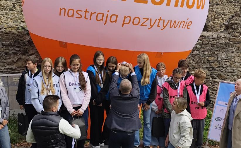 Trzy  czteroosobowe drużyny stoją na podium, Burmistrz dokonuje dekoracji medalowej. W tle mury obronne i duży balon z napisem Dzierżoniów.