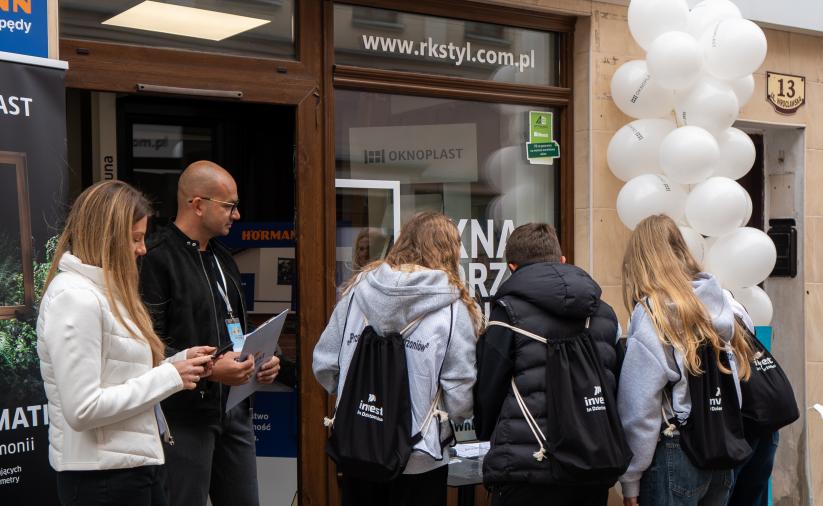 Uczestnicy gry terenowej stoją tyłem, pochyleni nad stołem przy stacji zadaniowej. Z lewej strony stoją opiekunowie stacji. W tle zarys budynku.