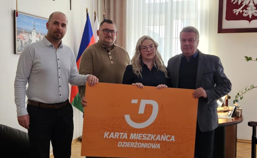 In the Mayor’s office, four people are standing: three men and one woman. All of them are jointly holding a large-format orange Dzierżoniów Resident Card.