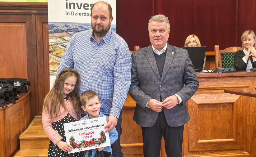 Laureat konkursu „Najładniejsza witryna świąteczna” z dziećmi trzymający dyplom za I miejsce, stojący obok burmistrza miasta podczas uroczystego wręczenia nagród w Sali Rycerskiej Urzędu Miasta.