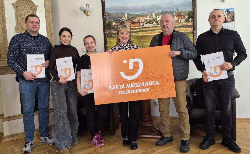 Grupa sześciu osób stoi w pomieszczeniu, wspólnie trzymając dużą, pomarańczową tablicę z napisem „Karta Mieszkańca Dzierżoniowa” oraz białym logotypem w kształcie litery „D”