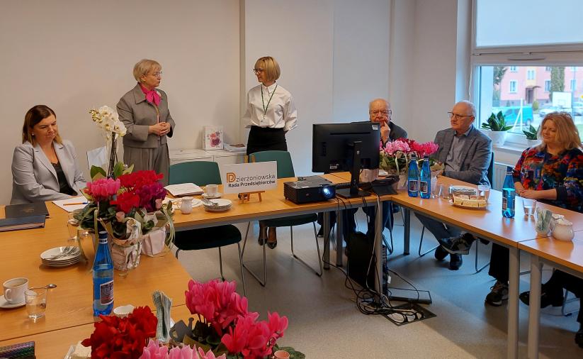Members of the Dzierżoniów Entrepreneurs’ Council at a conference table; two women talking in the center, with a projector, refreshments, and flowers in the background