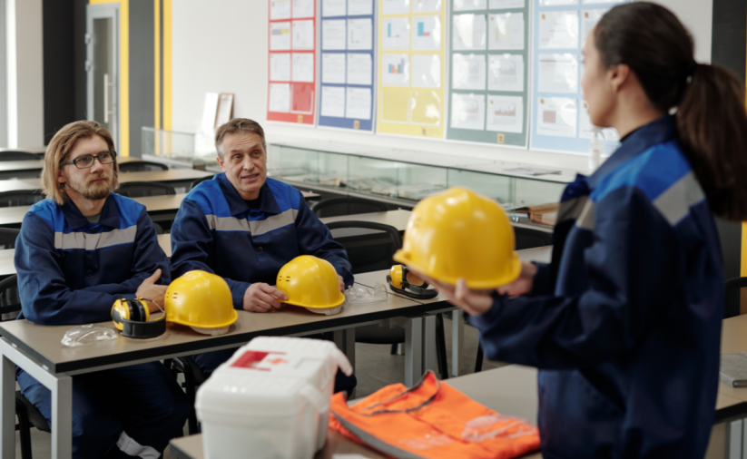 Kobieta w sali szkoleniowej trzyma żółty kask ochronny, naprzeciw niej siedzi dwóch mężczyzn w strojach roboczych. Na ławce leżą kaski, słuchawki i okulary ochronne oraz apteczka i kamizelka odblaskowa.