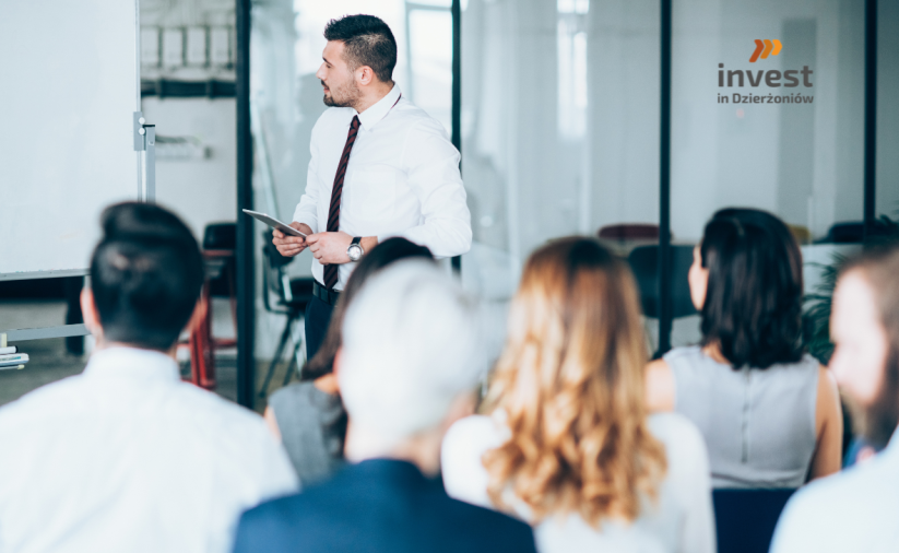 Mężczyzna prowadzi szkolenie w sali konferencyjnej, stojąc przy tablicy i trzymając tablet, a przed nim siedzi grupa uczestników; w tle widoczne logo „Invest in Dzierżoniów”