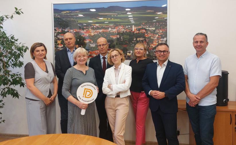 Grupa siedmiu osób w sali konferencyjnej na tle fotografii przedstawiającej Dzierżoniów. Jedna z osób trzyma okrągłe logo Dzierżoniowskiej Rady Przedsiębiorców.