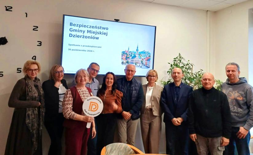 Grupa osób w sali konferencyjnej na tle monitora wyświetlajacego napis Bezpieczeństwo Gminy Miejskiej Dzierżoniów. Jedna z osób trzyma okrągłe logo Dzierżoniowskiej Rady Przedsiębiorców.