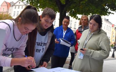 Drużyna pochyla się nad stołem i rozwiązuje zadanie na stacji gry. Obok stoi opiekun drużyny oraz organizatorzy stacji. W tle widoczny jest ratusz miejski w Dzierżoniowie