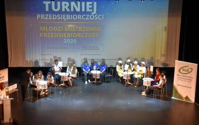Dziecie siedzą przy 5 stolikach na scenie, w tle slajd Międzyszkolny Turniej Przedsiębiorczosci