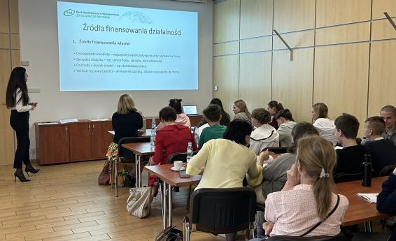 Sala wykładowa. Kobieta stoi przed wyświetlanym slajdem, a na sali siedzą uczniowie wraz opiekunami. 
