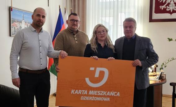 In the Mayor’s office, four people are standing: three men and one woman. All of them are jointly holding a large-format orange Dzierżoniów Resident Card.