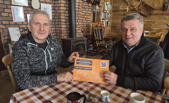 Dwóch mężczyzn przy stoliku trzyma naklejkę "Partner Programu. Tu honorujemy Kartę Mieszkańca Dzierżoniowa"