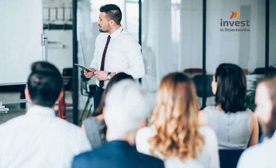 Mężczyzna prowadzi szkolenie w sali konferencyjnej, stojąc przy tablicy i trzymając tablet, a przed nim siedzi grupa uczestników; w tle widoczne logo „Invest in Dzierżoniów”