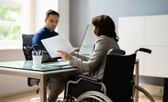 A woman in a wheelchair holds documents, while a man sits at a desk across from her.