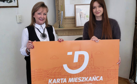 Dwie kobiety trzymają dużą, pomarańczową planszę z napisem Karta Mieszkańca Dzierżoniowa